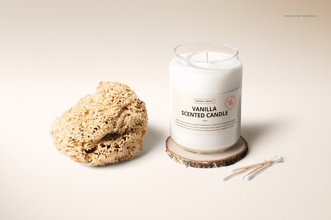 A white glass candle with a white label and the lettering "Vanilla scented candle" standing on a wooden stand, 4 cotton swabs and a stone.