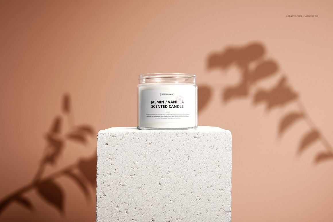 A white glass candle with a white label and the lettering "Jasmin/Vanilla scented candle" standing on a white stone block.