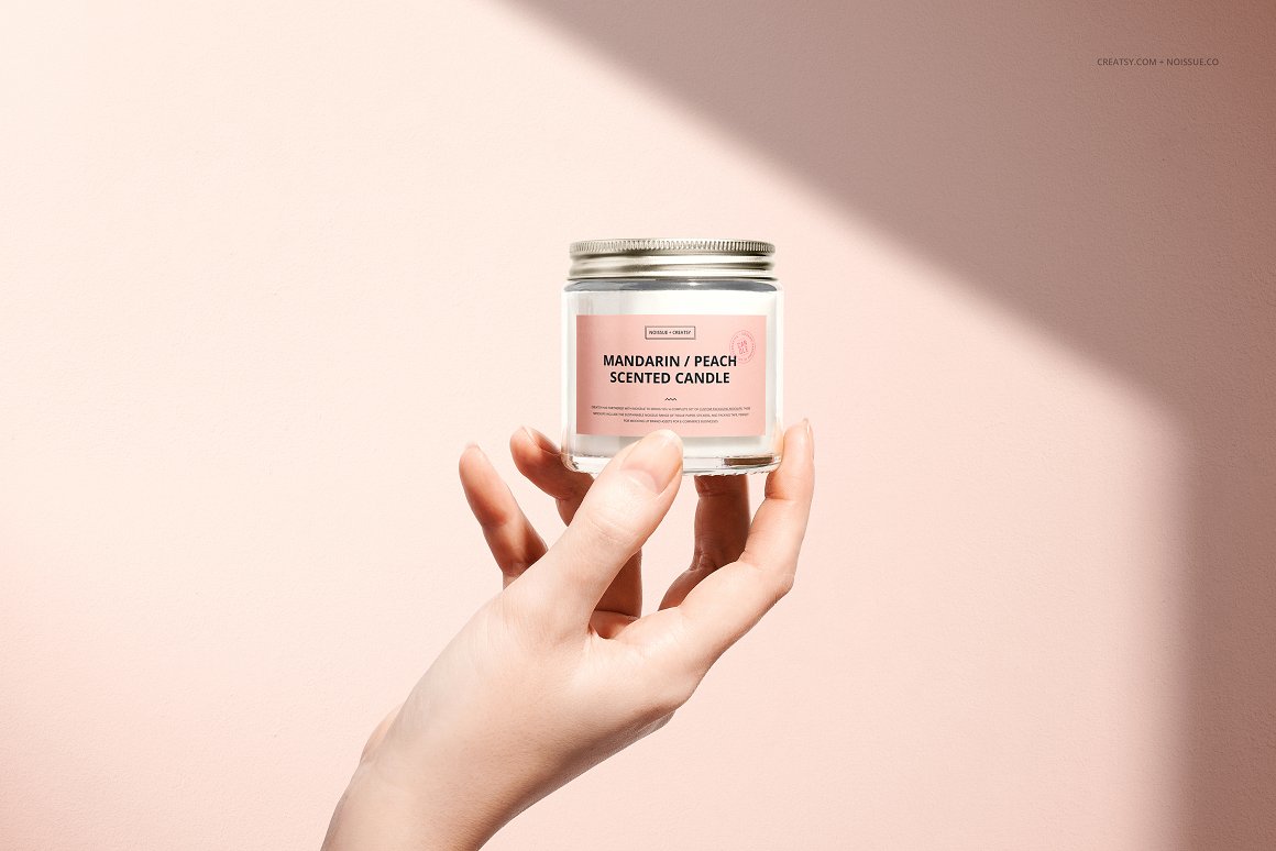 A woman's hand holding a white glass candle with a silver cap and a pink label with "Mandarin/Peach scented candle" written on it.