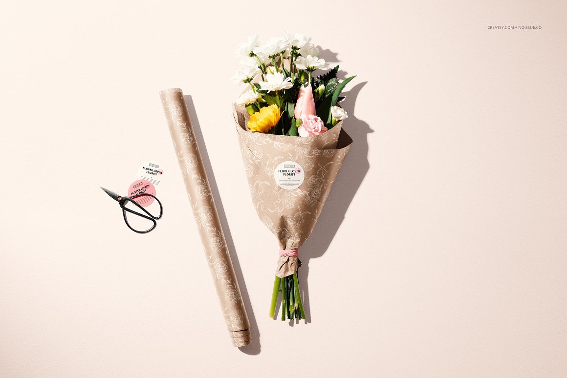 Bouquet of different flowers in beige wrapping paper with floral print with white label and the lettering "Flower Lover Florist", the paper itself and black scissors.