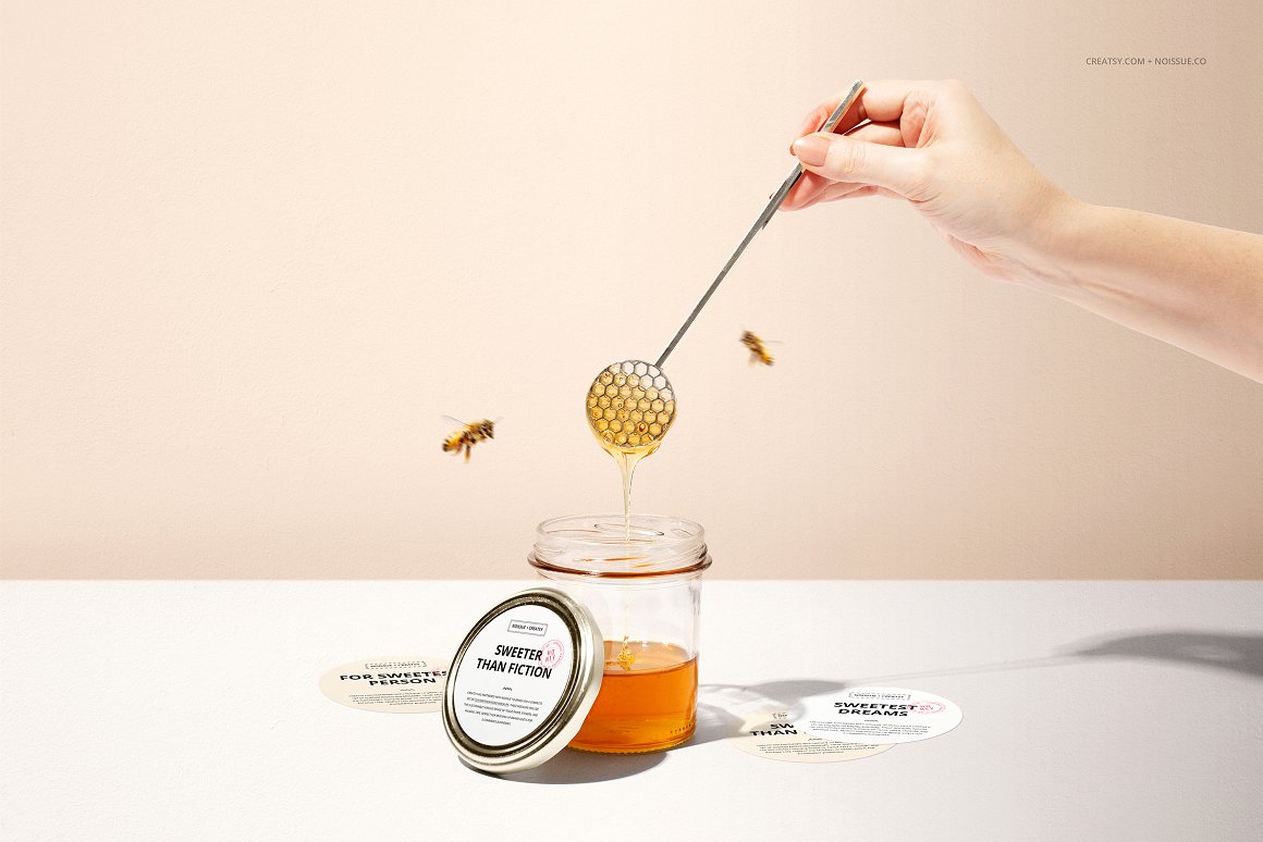 Glass jar with honey and a silver lid with a white label and the lettering "Sweeter than fiction" and 2 flying bees.