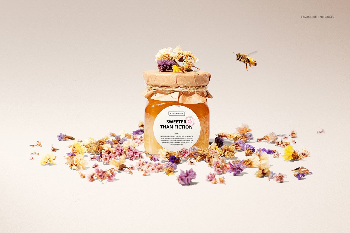 A glass jar with honey with a white label with the lettering "Sweeter than fiction" and a lid is packed in craft paper and wrapped with twine and dried flowers are scattered and flying bee.