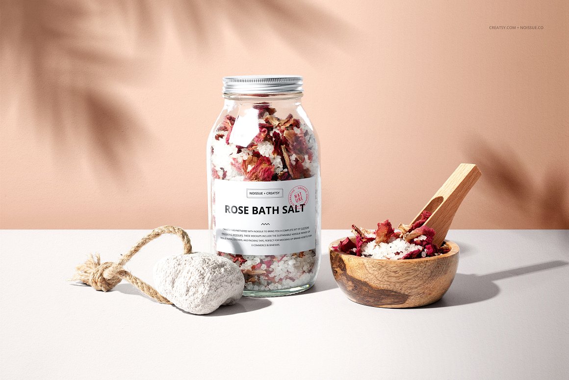 A glass jar with a silver lid and a white label with the lettering "Rose Bath Salt", a pumice stone and a wood bowl with a wood spoon containing rose bath salt.