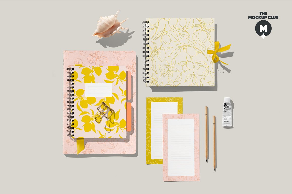 Pink spiral notebook mockup with lemon images, 2 beige pencils, pink notebook A4 mockup with fruit print, white spiral notebook with lemon images, shell, tube of paint and 2 cards on gray background.