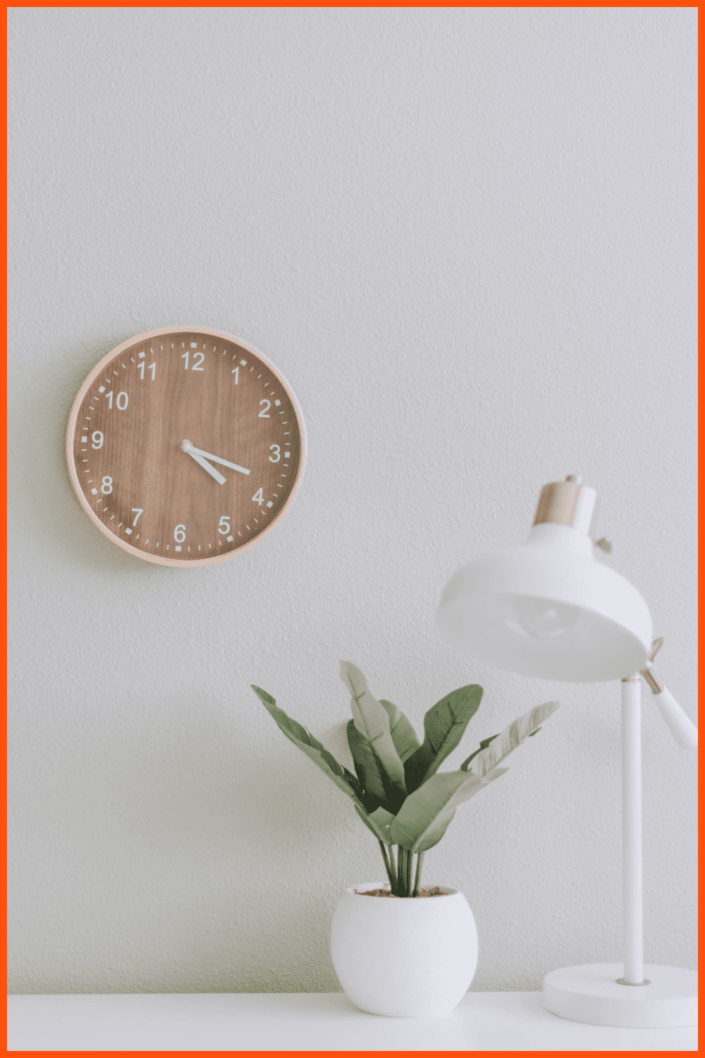 A clock on the wall, a white table lamp and a flower in a white pot on the table.