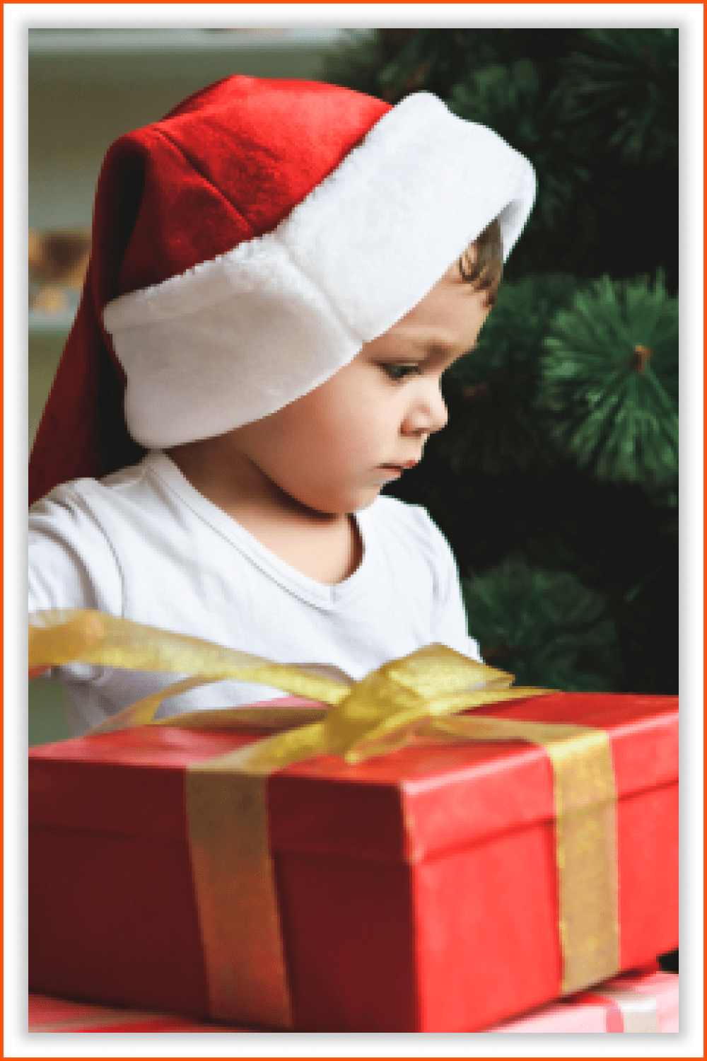 A small boy in a Santa hat with a big red gift and a green tree in the background.