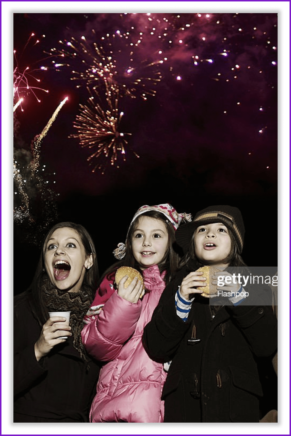 Mom and two girls with hamburgers watch fireworks in the dark sky.