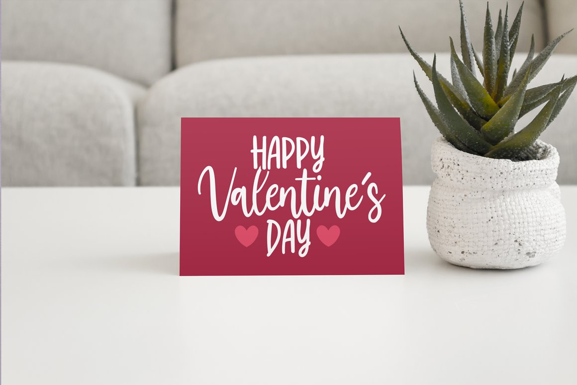 Pink greeting card with white lettering "Happy Valentine's Day" and 2 pink hearts on the table with aloe on a pot.