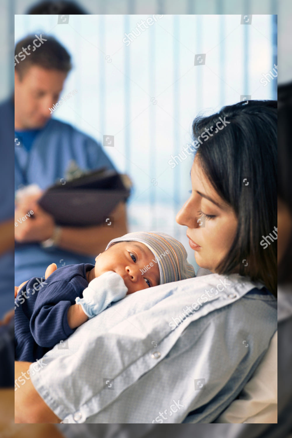 A side view of a mother holding a baby in the hospital bed while a doctor is sitting in the background checking reports.