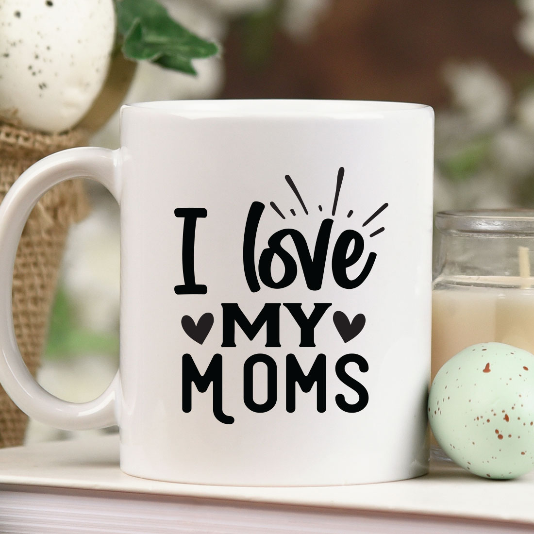 Coffee mug that says i love my moms next to a candle.