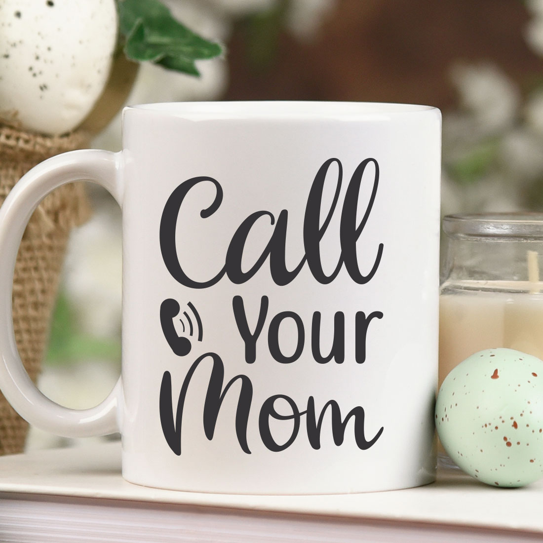 Coffee mug that says call your mom next to a candle.