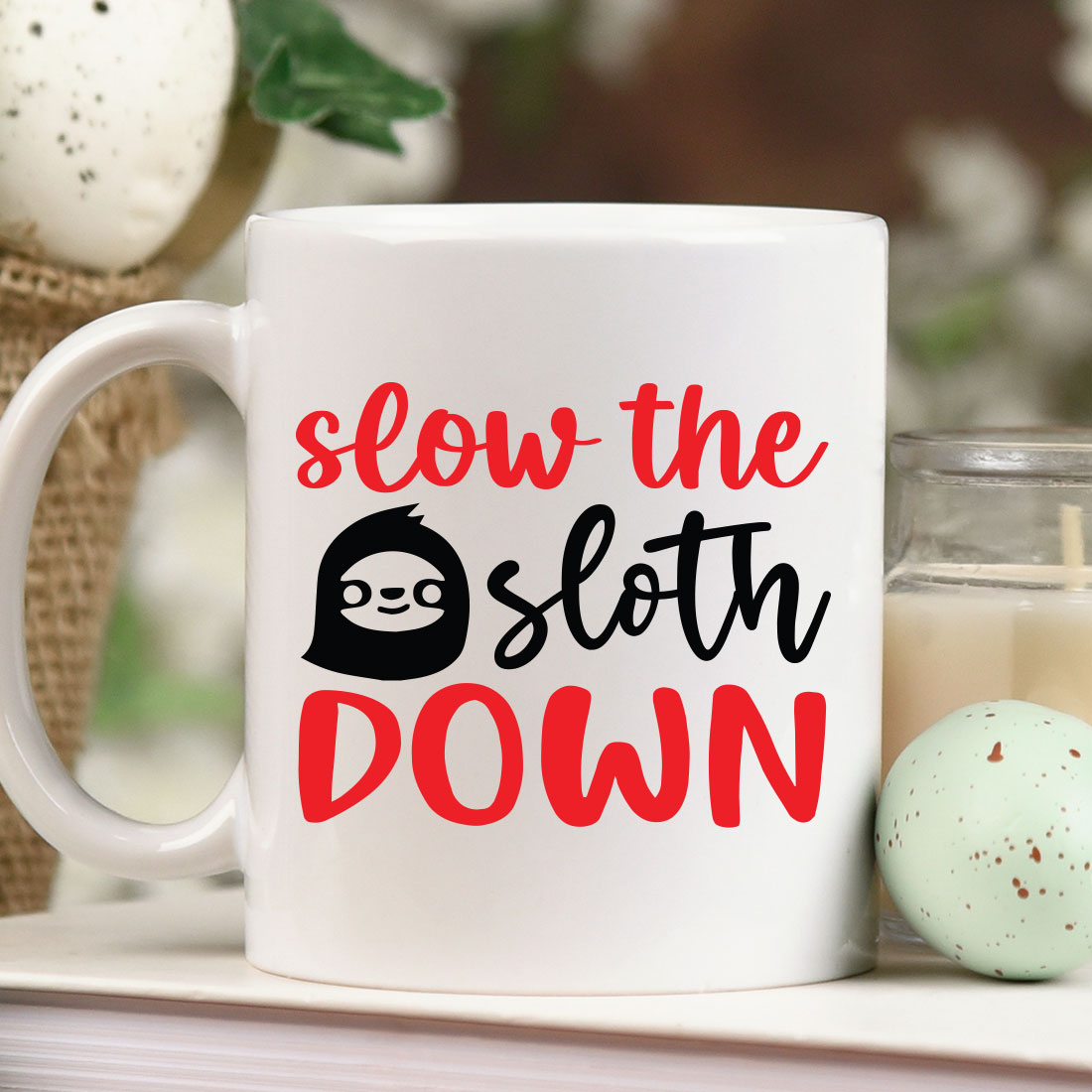 Coffee mug that says slow the sloth down next to a candle.