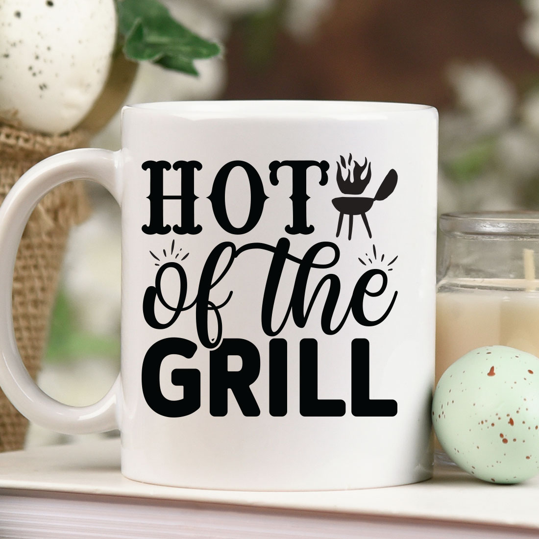 Coffee mug that says hot of the grill next to a candle.