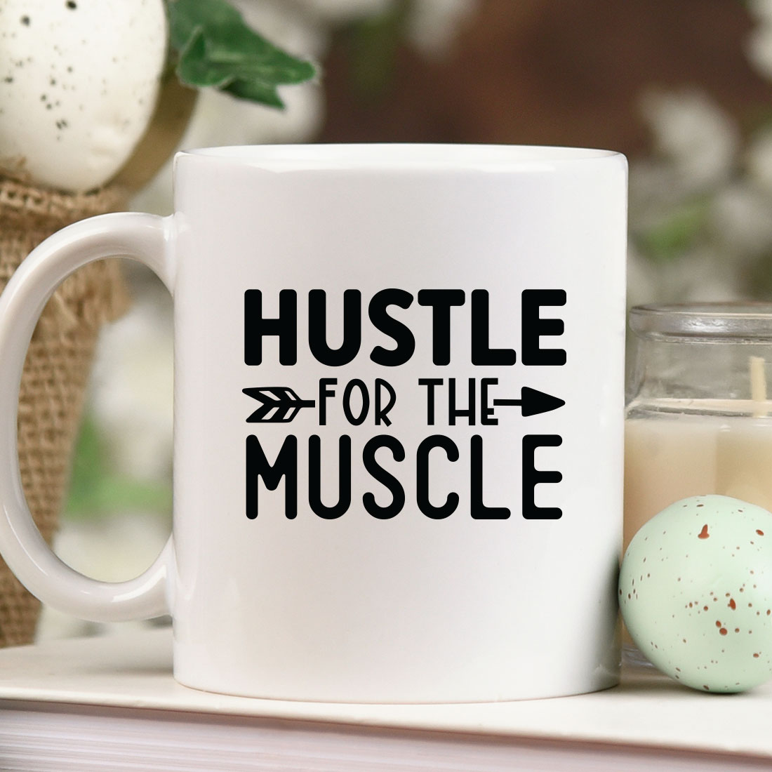 Coffee mug that says hustle for the muscle next to a candle.