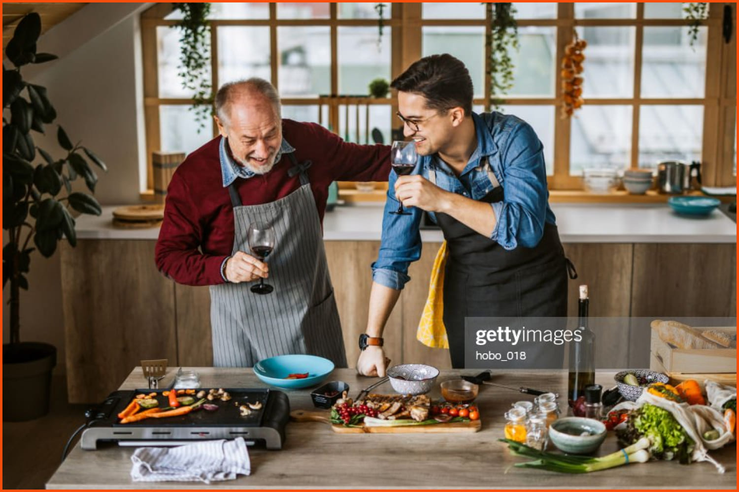 Senior father and young adult son preparing family lunch together in domestic home kitchen. Drink wine and have fun.