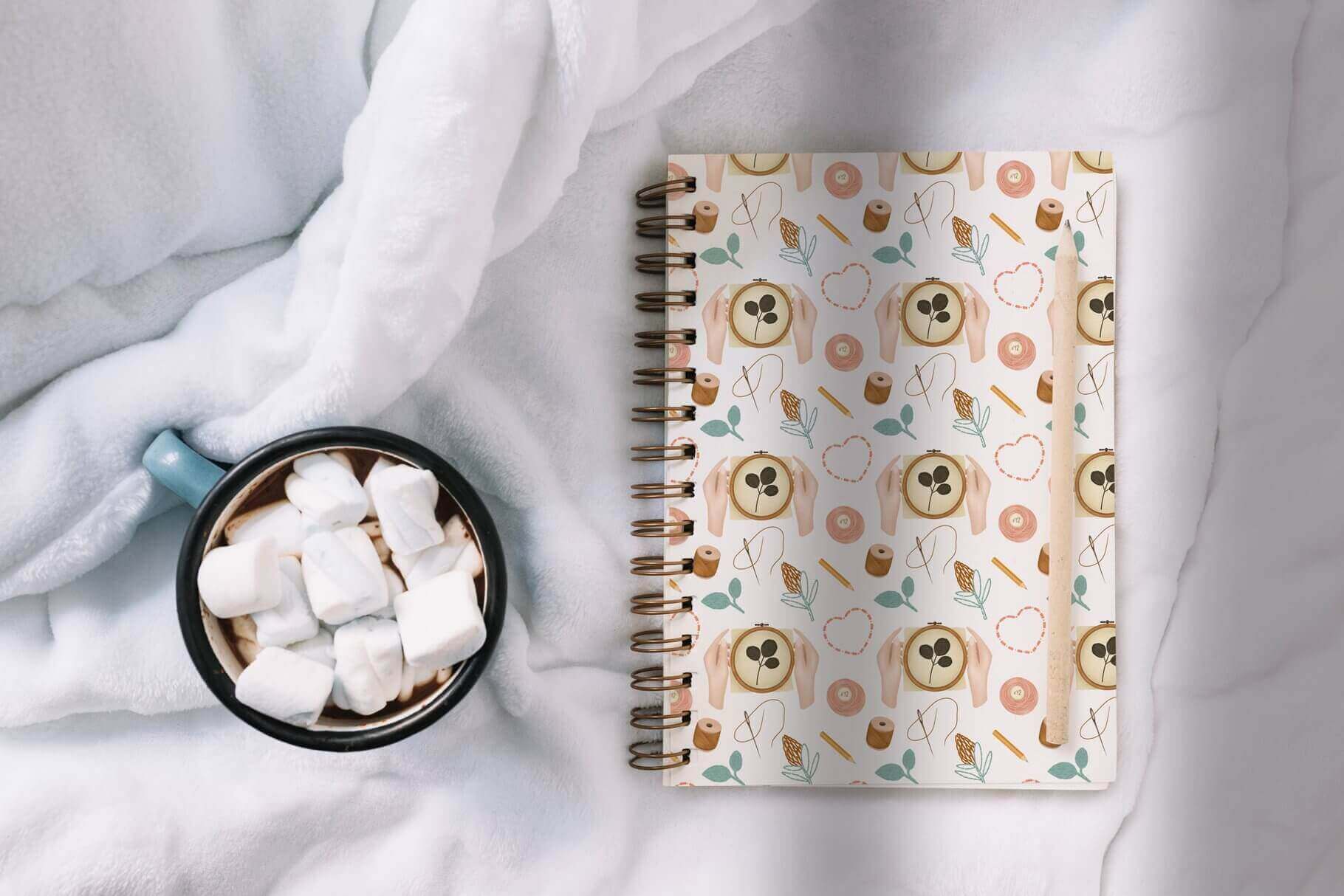 On a soft bedspread is a cup of marshmallows and a notebook with a handicraft print.